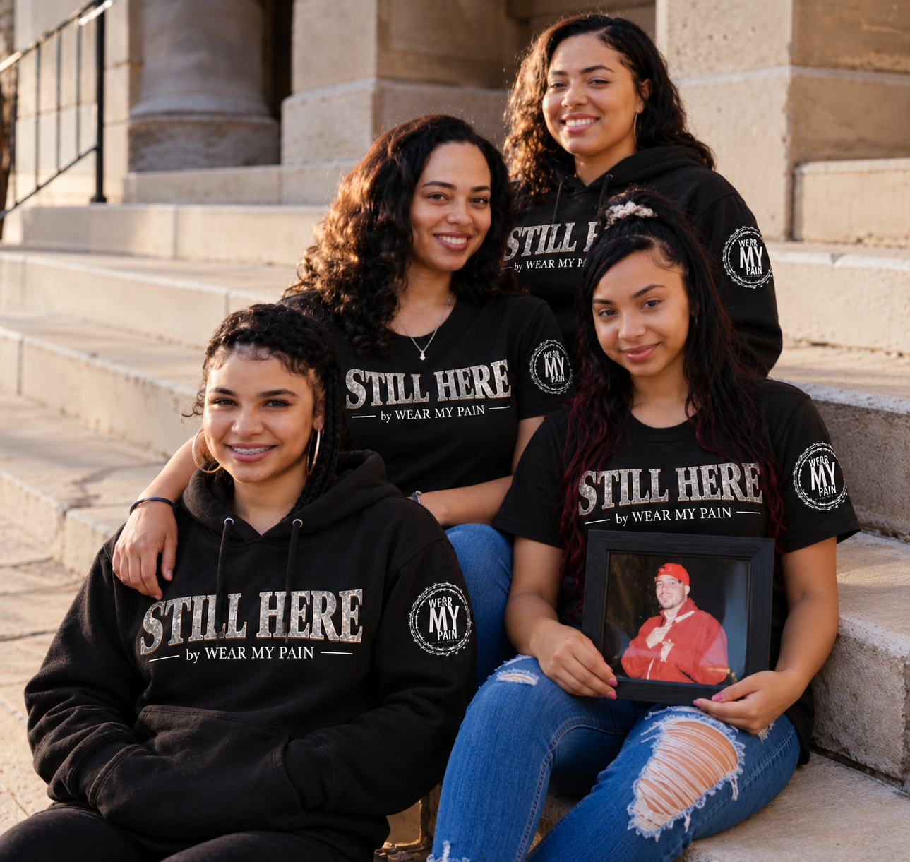 Four women wearing STILL HERE by Wear My Pain apparel sitting together on stone steps, one holding a framed photograph of a loved one